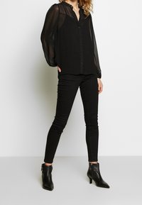 Mustapaitainen läpinäkyvä paita, jossa on röyhelökaulus ja nappi-etuosa, yhdistetty mustiin slim fit -housuihin ja nilkkureihin. Sujuva tekstuuri kauttaaltaan.