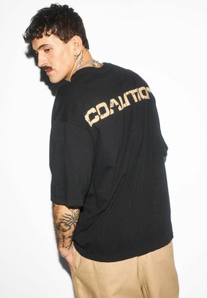 Mężczyzna z krótkimi kręconymi włosami i tatuażami, ubrany w czarny oversize'owy T-shirt z napisem "COALITION" na plecach oraz beżowe spodnie.