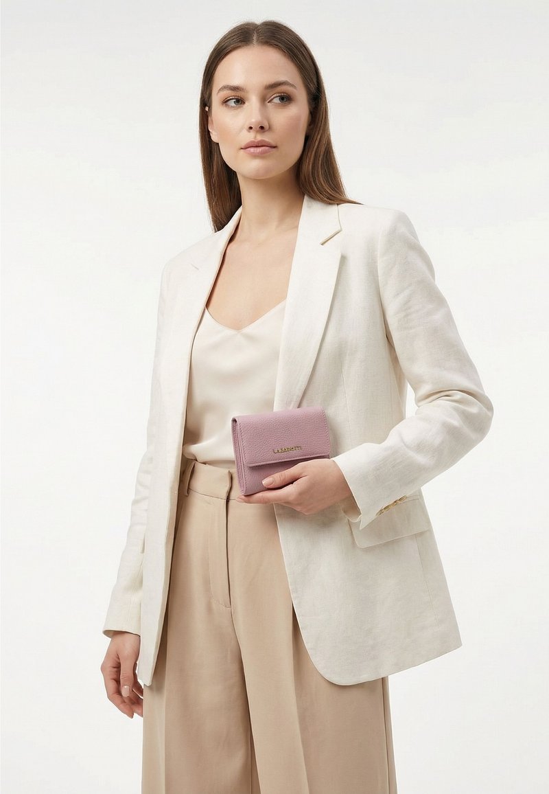 Frau in beiger Hose und weißem Blazer hält eine kleine rosa Geldbörse mit goldener Schrift vor einfachem Hintergrund.