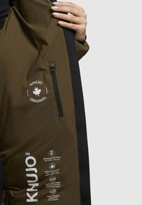 Brun og sort vandtæt jakke med en lynlås lomme, KHUJO-logo og plejeikoner for stof. Har et let design til varme og vindbeskyttelse.
