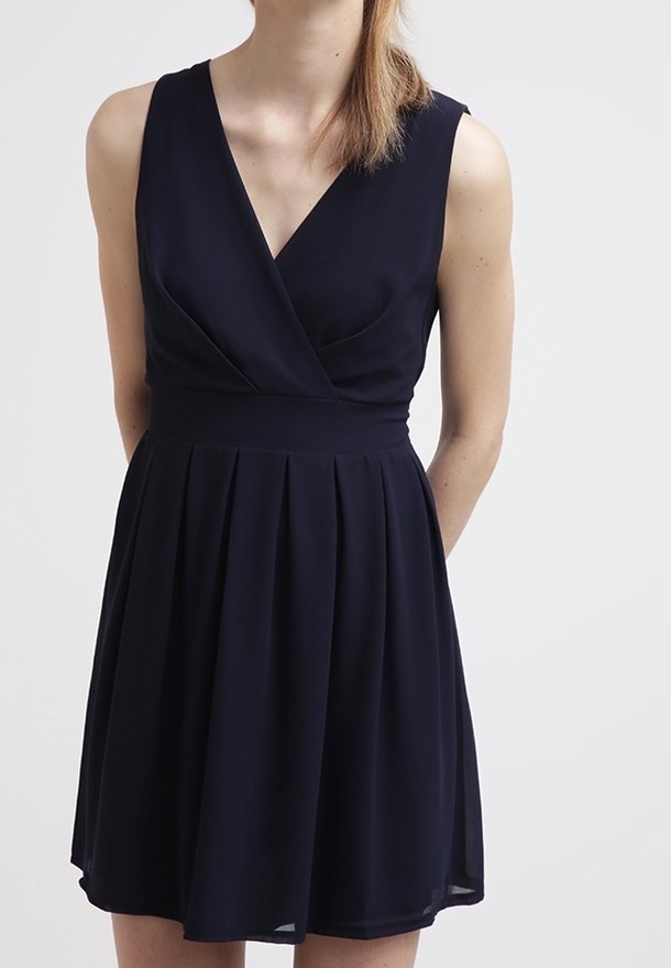 Femme portant une robe noire sans manches, longueur genoux, avec un col en V et une jupe plissée, debout devant un fond blanc uni.