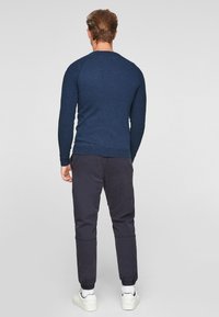 Dunkelblauer Pullover mit langen Ärmeln und geripptem Saum, kombiniert mit dunklen Hosen; weiße Sneaker sichtbar. Einfaches, tailliertes Design.