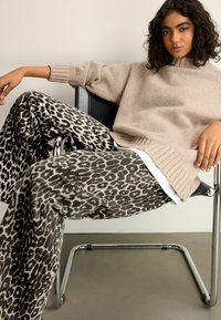Béžový oversized pletený sveter s rebrovanými manžetami v kombinácii s čiernymi a šedými nohavicami s leopardím vzorom a širokými nohavicami, sedením na chrómovej stoličke.