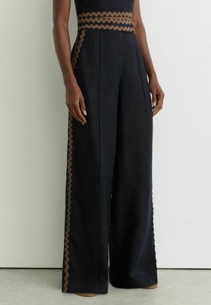 Lange schwarze Hosen mit weitem Bein, die braune wellenförmige Verzierungen entlang der Taille und der Außenseiten aufweisen, getragen von einer stehenden Person vor einer schlichten Wand.