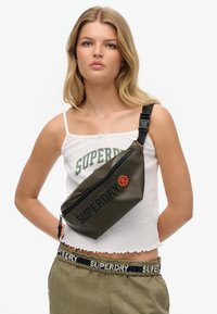 Oliwkowa saszetka na pas z zamkiem błyskawicznym, z wyrazistym czarnym logo "SUPERDRY" i czerwonym akcentem. Wykonana z trwałego, gładkiego materiału.