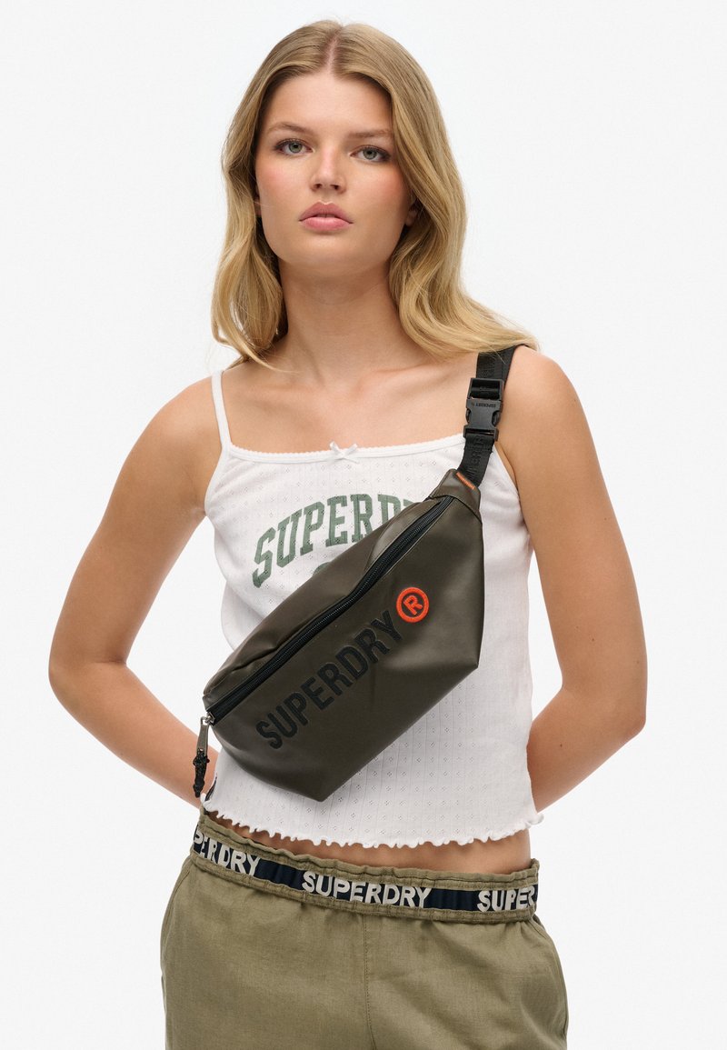Oliwkowa saszetka na pas z zamkiem błyskawicznym, z wyrazistym czarnym logo "SUPERDRY" i czerwonym akcentem. Wykonana z trwałego, gładkiego materiału.
