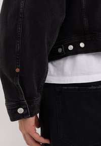 Sort denimjakke med en let oversized pasform, prydet med sølvfarvede trykknapper og et struktureret stof. Beklædningen er lagdelt med en hvid skjorte.
