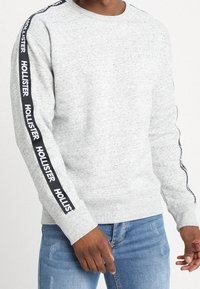 Man som bär en ljusgrå rundhalsad sweatshirt med svart tejp som har texten "Hollister" längs ärmarna, i kombination med blå jeans.