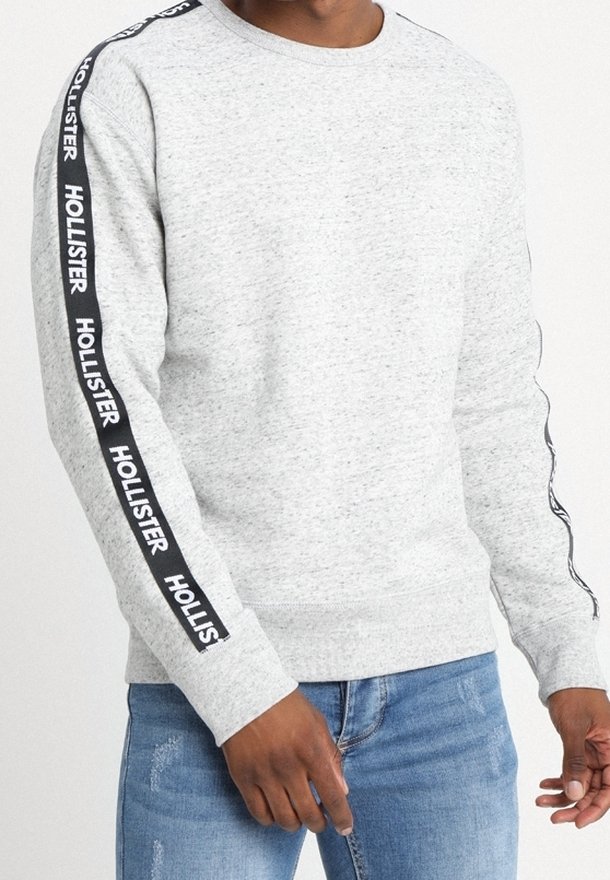 Man som bär en ljusgrå rundhalsad sweatshirt med svart tejp som har texten "Hollister" längs ärmarna, i kombination med blå jeans.