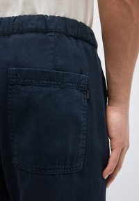 Κοντινή λήψη του χεριού ενός ατόμου δίπλα στην πίσω τσέπη σκούρων μπλε παντελονιών με ελαστική μέση, που διαθέτουν μια μικρή ετικέτα στη ραφή της τσέπης.