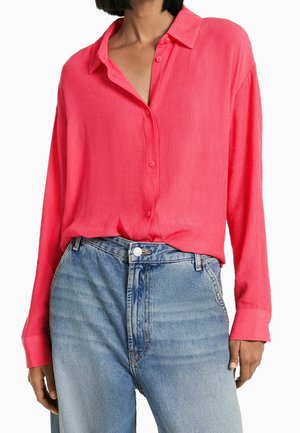 Chemise à manches longues sans col de couleur rose vif, présentant un tissu texturé et des boutons à l'avant, associée à un jean clair taille haute.