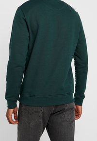 Dark green sweatshirt with long sleeves, a round neck, and ribbed cuffs and hem. Fabric appears soft, with minimal stitching detail.
