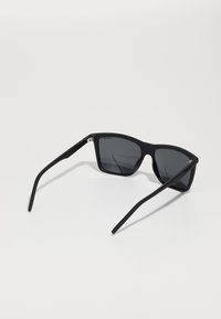 Ochelari de soare negri cu finisaj mat, cadru unghiular, lentile întunecate și temple drepte, elegante. Branding minimalist aproape de balama.
