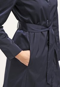 Femme portant un trench-coat ceinturé bleu marine foncé avec des boutons, main dans la poche latérale, vue de la moitié du torse jusqu'à la hanche.