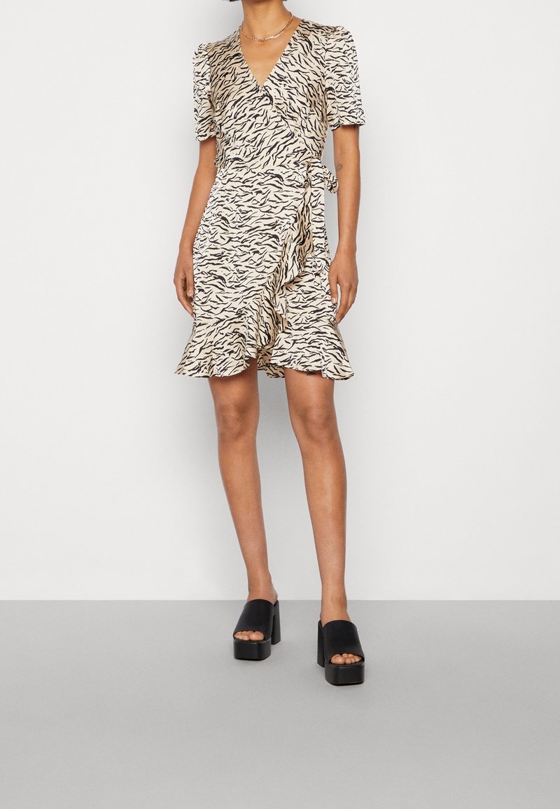 Femme portant une robe portefeuille beige avec imprimé zèbre noir et ourlet à volants, associée à des sandales plateformes noires.