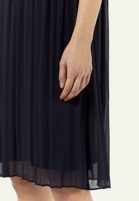 Granatowa sukienka z plisami o przezroczystej fakturze, sięgająca do połowy łydki. Ręka delikatnie trzyma materiał u dołu, ukazując płynny ruch.