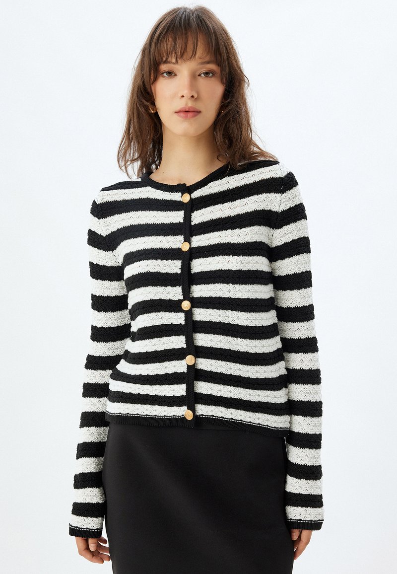 Randig cardigan i svart och vitt med texturerat stickat, rund halsringning och guldknappar, i kombination med en svart kjol.