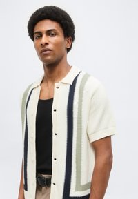 Άνδρας με κοντά σγουρά μαλλιά που φοράει λευκό πλεκτό μπλουζάκι με κοντά μανίκια και μαύρες και γκρι κάθετες ρίγες πάνω από μαύρο τοπ.