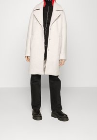 Ljusgrå ullrock med slagkrage, två framknappar och stora fickor; bärs över en svart polotröja och svarta byxor.