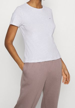 Kobieta w jasnoszarej obcisłej koszulce z krótkim rękawem i luźnych, purpurowo-różowych spodniach, stojąca z jedną ręką w kieszeni na jednolitym tle.