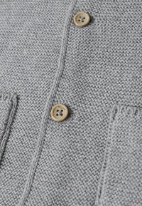 Grau gestrickte Strickjacke aus strukturiertem Stoff mit zwei beigen Knöpfen und zwei vorderen Taschen. Betonte Nähte entlang der Kanten.