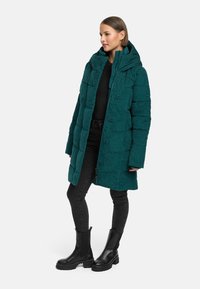 Teal puffjacka med huva, med en kviltad design, tryckknappar och snedställda sidofickor, kombinerad med svarta skinny jeans och stövlar.