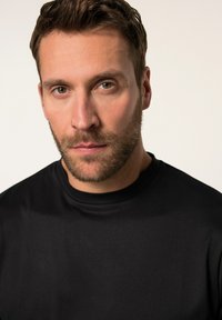 Camisa negra de manga corta con un escote redondo, hecha de una tela suave; vestida por un hombre con el cabello corto y barba, de frente.
