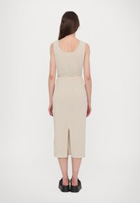 Rochie midi tricotată de culoare bej deschis, cu textură canelată, design fără mâneci, slit la spate și talie ajustată. Pur și simplu cu pantofi negri masivi.