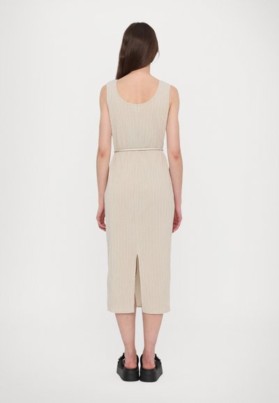 Vestido midi de malha em bege claro com textura em canelado, design sem mangas, fenda nas costas e cintura ajustada. Usado com sapatos pretos robustos.