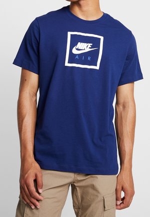 Mężczyzna ubrany w niebieską koszulkę Nike Air i beżowe spodnie typu cargo, stojący z rękami luźno opuszczonymi wzdłuż ciała na jednolitym tle.