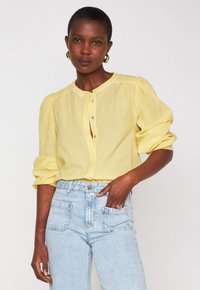 Frau mit kurzem Haar, die goldene Creolen trägt, eine gelbe Bluse mit Knöpfen und hochgekrempelten Ärmeln, helle blaue hochtaillierte Jeans und einen pinken Ring.