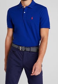 Kungsblå polo med krage och tre knappar, har en delvis röd broderad logotyp på bröstet, tillsammans med ett texturerat grått bälte.