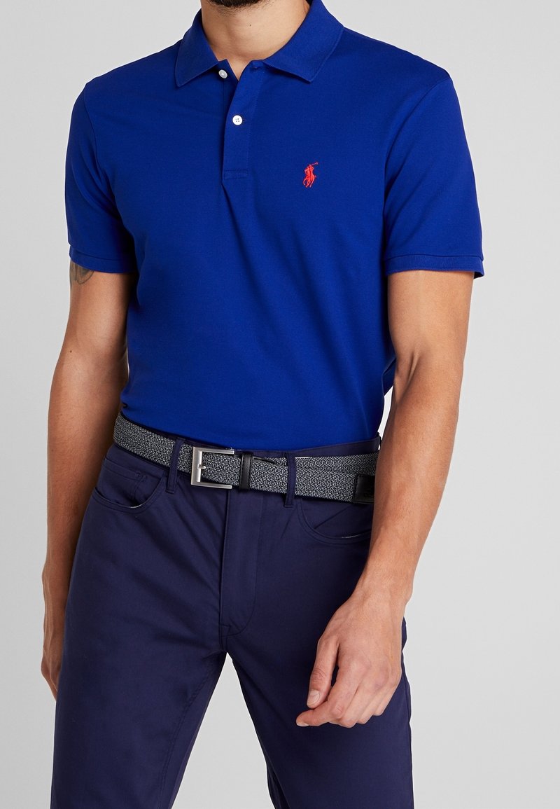 Kungsblå polo med krage och tre knappar, har en delvis röd broderad logotyp på bröstet, tillsammans med ett texturerat grått bälte.