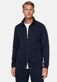 Giacca a zip blu navy in tessuto morbido, caratterizzata da un colletto alto, tasche laterali, polsini a coste e una semplice chiusura con zip. Strato interno bianco visibile.
