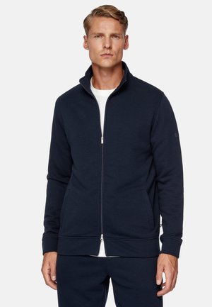 Navy zip-up jacket in soft fabric, featuring a high collar, side pockets, ribbed cuffs, and a simple zipper closure. White inner layer visible.