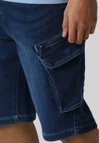 Mørkeblå denimshorts med en tætsiddende pasform. Har cargo-lommer med lynlåse og kontrastfarvet syninger langs sømmene.