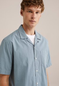 Camicia a maniche corte con bottoni di colore azzurro chiaro, con righe bianche verticali, caratterizzata da una tasca sul petto e un classico colletto.