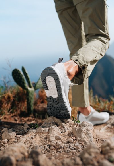 Scarpa sportiva bianca con suola grigia testurizzata, dotata di un tassello per il sollevamento e pannelli in mesh, posizionata su un terreno roccioso con vegetazione nelle vicinanze.