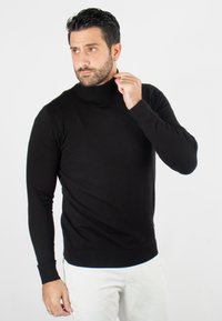Un pull col roulé noir en tissu doux, au design ajusté avec de longues manches et des poignets côtelés, porté avec un pantalon de couleur claire.