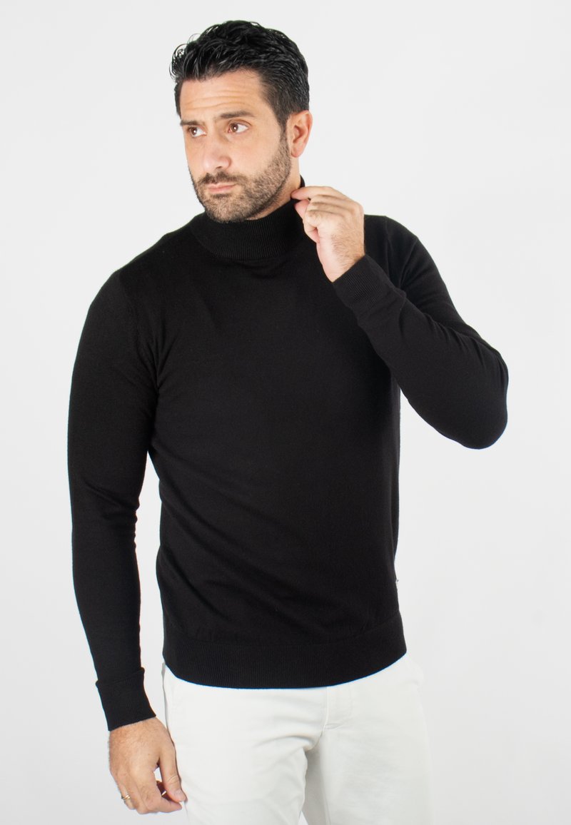 Un pull col roulé noir en tissu doux, au design ajusté avec de longues manches et des poignets côtelés, porté avec un pantalon de couleur claire.