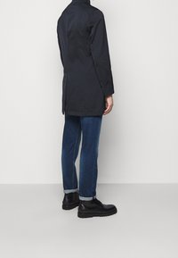 Černý kabát s vysokým límcem, slim fit a rovnými zády. Nositelný přes modré džíny a černé boty, zdůrazňující elegantní a moderní siluetu.