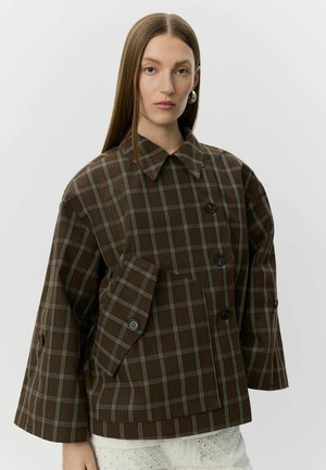 Donna con capelli lunghi e lisci indossa una giacca oversize marrone a quadri con grandi bottoni e maniche larghe sopra pantaloni bianchi strutturati.