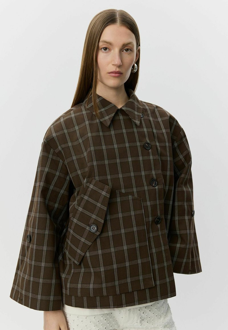 Femme aux longs cheveux raides porte une veste surdimensionnée à carreaux marron avec de gros boutons et de larges manches sur un pantalon blanc texturé.