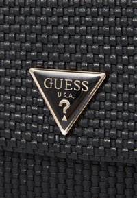 Schwarze gewebte Textur mit einem dreieckigen GUESS-Logo in Gold, das "U.S.A." und ein Fragezeichen-Design in der Mitte zeigt.