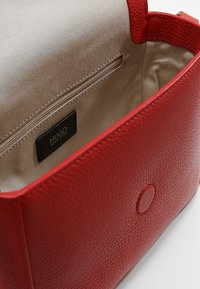 Rød læderhandtaske med en struktureret overflade, der har en beige indvendig side, lynlås lomme og et sort mærke. Rund knaplukning som detalje.