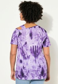 Fialové tričko s potiskem tie-dye a krátkým rukávem, s uvolněným střihem a výřezy na rameni. Materiál vypadá měkký a má texturovaný pocit.