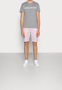 Camiseta de algodón gris con "CALVIN KLEIN JEANS" impreso en blanco, combinada con pantalones cortos rosa claro y zapatillas blancas. Conjunto simple y casual.