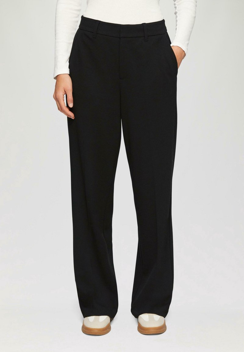 Pantalon noir à jambes droites porté avec un haut blanc à manches longues et des baskets beige avec des accents blancs, mains reposant sur les côtés et dans la poche.