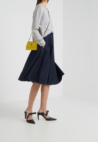 Un pull gris, une jupe évasée bleu marine, des chaussures à talons hauts noires avec des accessoires ronds, et un petit sac en bandoulière jaune.