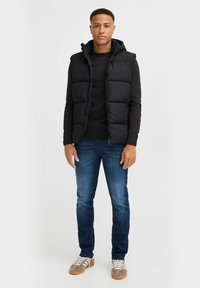 Schwarze Steppweste mit Kapuze über einem schwarzen Sweater, kombiniert mit dunkelblauen Jeans und grauen Sneakern mit weißen Streifen an der Seite.